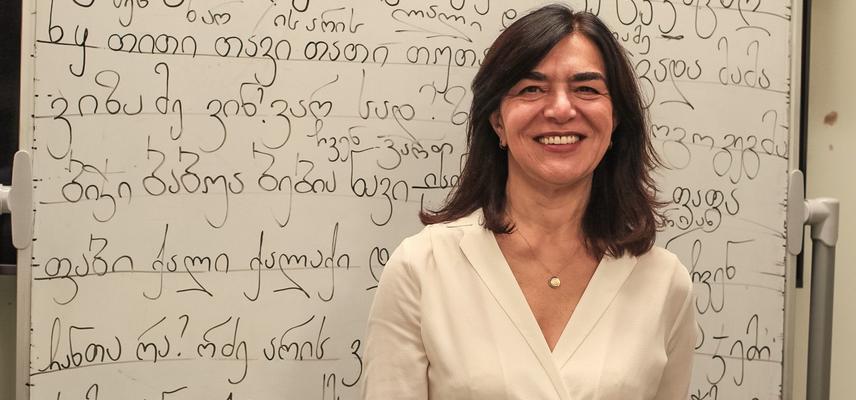 Lia Chokoshvili has shoulder-length dark hair. She is wearing a white v-neck blouse and standing in front of a whiteboard covered in Georgian script.