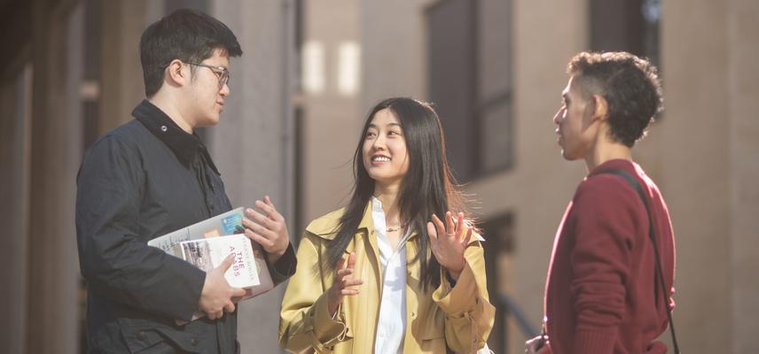 Three students standing outside chatting. They are wearing light jackets and two of them are holding books.