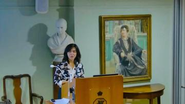 Lia Chokoshvili is standing and speaking behind a brown lectern. She is wearing a black and white patterned blazer
