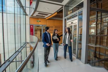 Three students walking through a corridor of a modern department building, surrounded by large glass windows. 