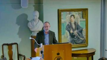 A man with short grey hair and a grey beard presenting a lectern. 