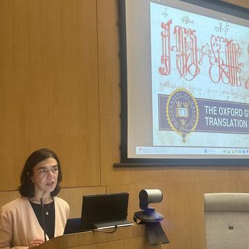 Lia Chokoshvili has shoulder-length brown hair. She is standing at a wooden lectern, with a presentation on a large screen behind her.