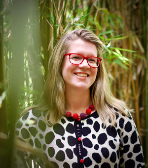 Kate has blonde shoulder length hair and red glasses. She's wearing a cardigan with a white background with a black pattern and red buttons.