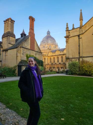 ellie has long dark brown hair worn in a pony tail. She's wearing a navy blue jacket with long royal blue scarf. She's standing in a college garden with college buildings and the Bodleian Library behind her.