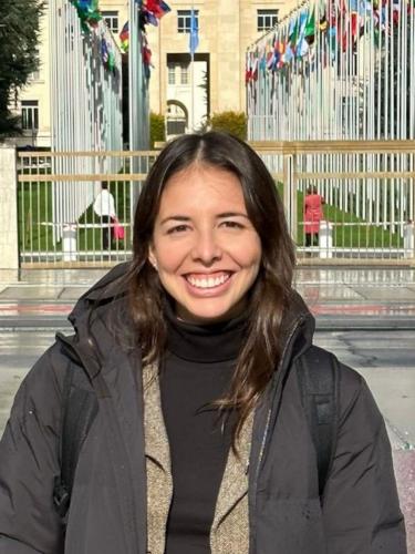 luciana has long dark brown hair and is smiling at the camera. She's wearing a black jumper and grey woollen cardigan under a grey anorak. She's standing in front of the country flags outside the UN Palais des Nations, Geneva.