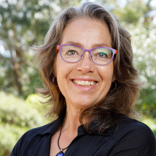Rita Abrahamsen has brown shoulder-length hair and wearing purple glasses. She's wearing a navy blue top and smiling at the camera 