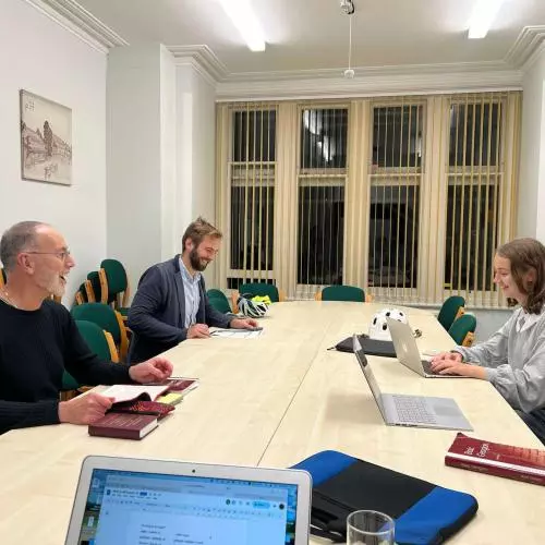 Three students sitting around a long table, looking at laptops and papers while smiling. 