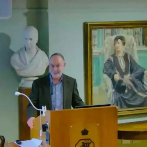 A man with short grey hair and a grey beard presenting a lectern. 
