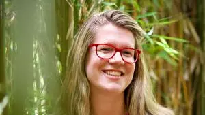 Kate has blonde shoulder length hair and red glasses. She's wearing a cardigan with a white background with a black pattern and red buttons.