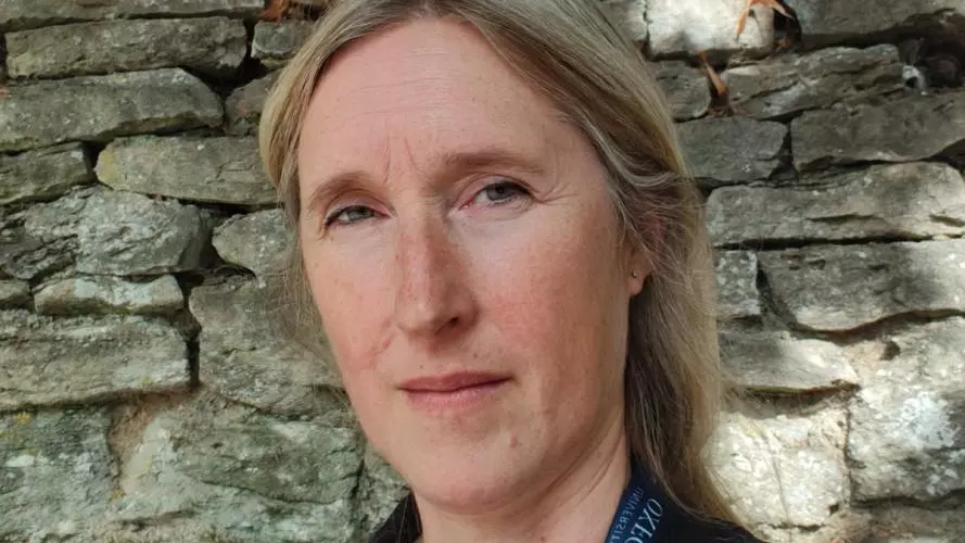 Dr Eklund has long blond hair and is wearing a dark blazer, standing in front of an old brick wall