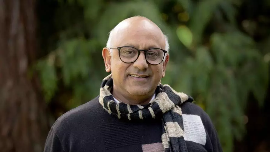 Premesh has grey hair, dark rimmed glasses and is wearing a blue and grey scarf over a navy blue sweater.  There is a blurred green background resembling trees behind him. 