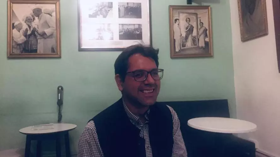 Adnan has short dark brown hair, is wearing glasses and smiling broadly at the camera. He's wearing a blue and white gingham shirt under a navy blue woollen waist coat. Behind him is a pale green painted wall with framed black and white photos of people.   