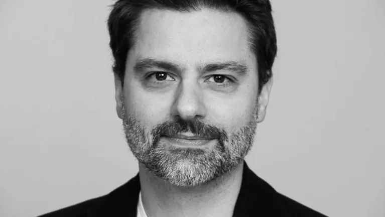 A black and white photo of Felipe who has short dark hair and grey beard. He's wearing a white v neck t shirt under a smart black jacket. 