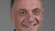 Othon has short grey hair and smiling broadly at the camera. He's wearing a sky blue shirt under a dark blue jumper 