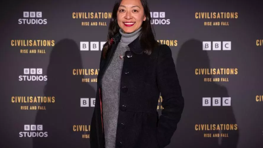 Jessica has long dark hair and is smiling at the camera. She's wearing a grey dress under a long black coat. She's standing against a 'BBC Civilisations: Rise and Fall' backdrop