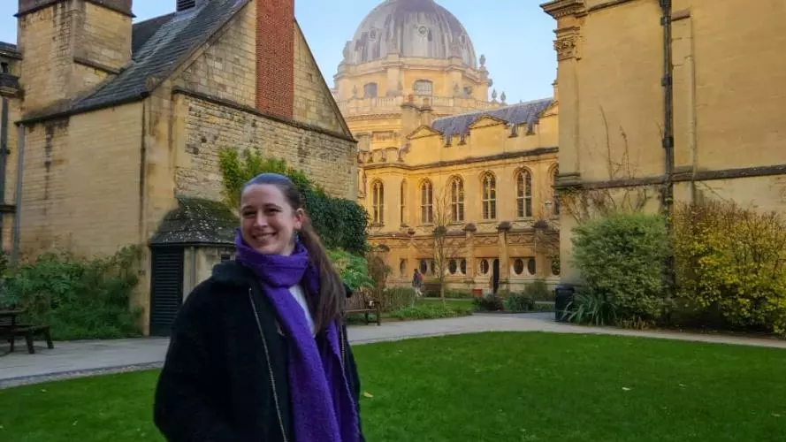 ellie has long dark brown hair worn in a pony tail. She's wearing a navy blue jacket with long royal blue scarf. She's standing in a college garden with college buildings and the Bodleian Library behind her.