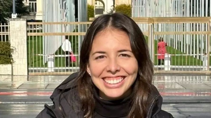 luciana has long dark brown hair and is smiling at the camera. She's wearing a black jumper and grey woollen cardigan under a grey anorak. She's standing in front of the country flags outside the UN Palais des Nations, Geneva.