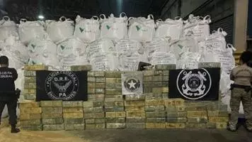 A wall of white industrial bulk bags stacked in rows behind neatly piled brown wrapped packages. Two armed officers are standing facing the packages.