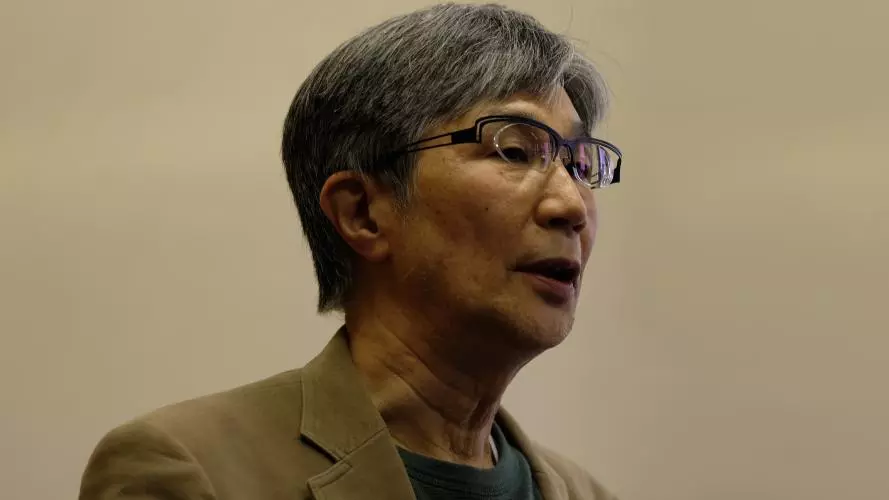 takehiko kariya has short grey hair, glasses and is wearing a grey top under a tan coloured jacket. 