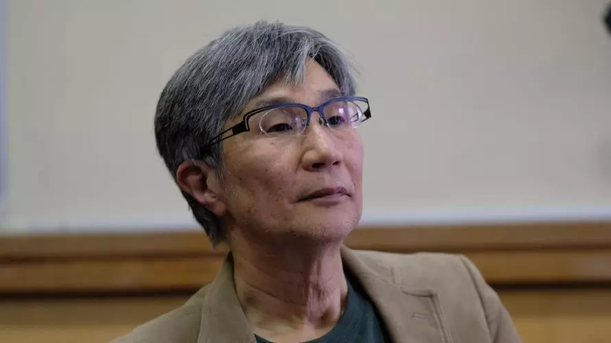 takehiko kariya has short grey hair, glasses and is wearing a grey top under a tan coloured jacket