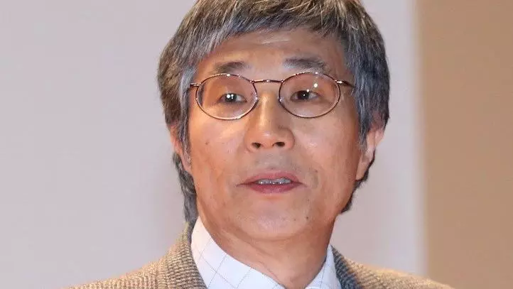 takehiko kariya has short grey hair, wearing glasses, and dressed in a white shirt, black and white striped tie, and brown woollen jacket