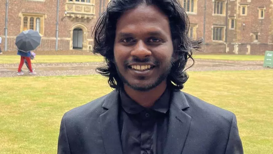 Roya has black wavey hair and a bead. He's dressed in a dark grey shirt and jacket as is standing in a college quad.