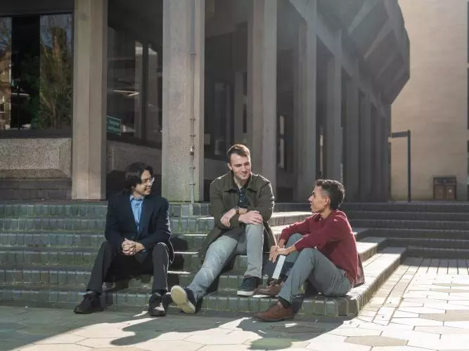 Three students sitting on steps outside a building with large grey pillars.