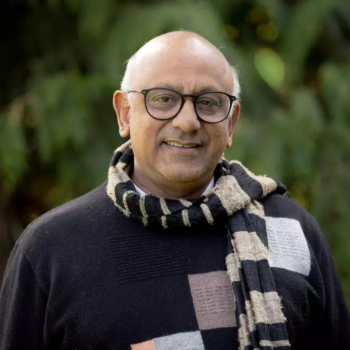 Premesh has grey hair, dark rimmed glasses and is wearing a blue and grey scarf over a navy blue sweater.  There is a blurred green background resembling trees behind him. 
