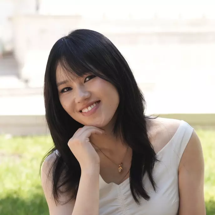  Mingdan has long black hair, is resting her chin on her hand, and is smiling at the camera. She's wearing a sleeveless white top