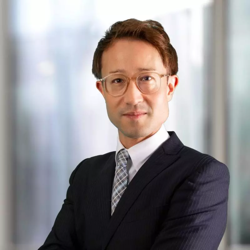 Dr Buchmeier has short brown hair and is wearing clear-rimmed glasses. He is wearing a pinstripe suit, shirt and tie. He is standing with his arms folded, looking at the camera. 