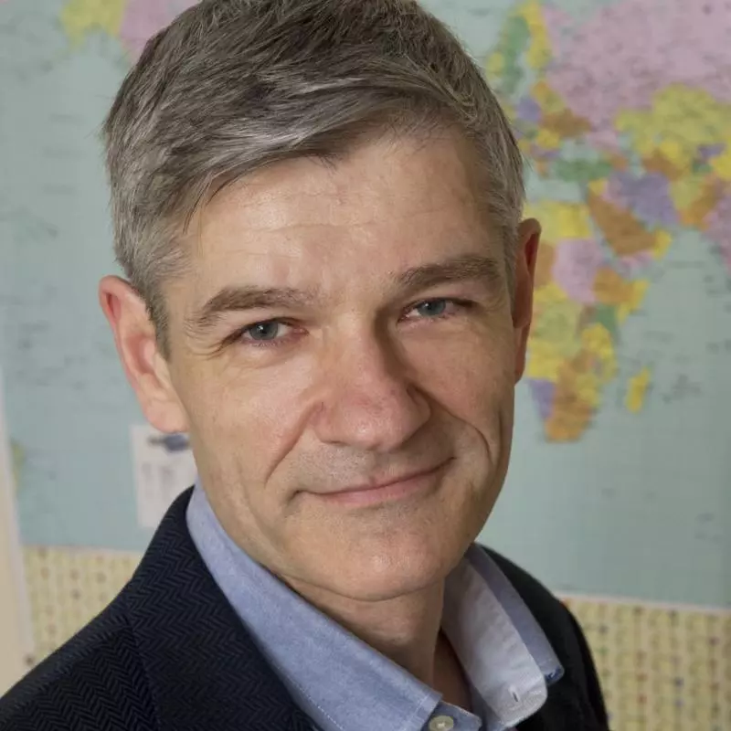 Roy Allison has short grey hair and is looking towards the camera. He's wearing a pale blue shirt with smart navy blue jacket. There is a world map behind him. 