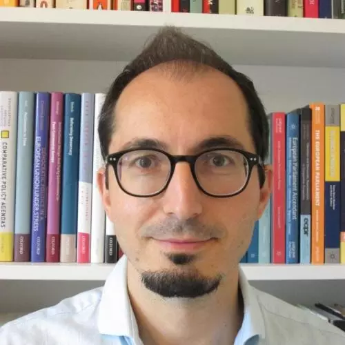 Dr Chiru has brown hair and a short beard. He is wearing dark round glasses and standing in front of a bookshelf