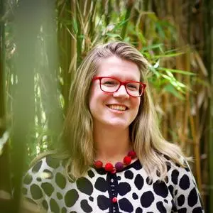 Kate has blonde shoulder length hair and red glasses. She's wearing a cardigan with a white background with a black pattern and red buttons.