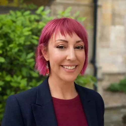 Janey has pink hair in a short bob with fringe. She's wearing a wine red top with navy blue jacket