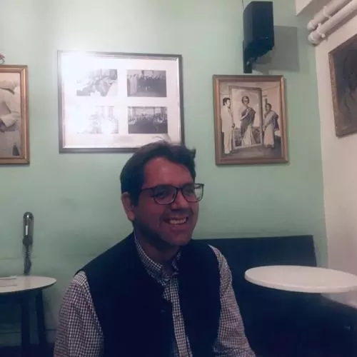 Adnan has short dark brown hair, is wearing glasses and smiling broadly at the camera. He's wearing a blue and white gingham shirt under a navy blue woollen waist coat. Behind him is a pale green painted wall with framed black and white photos of people.   