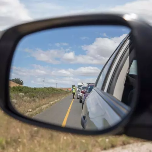 A close-up of a car wing mirror, which is reflecting a queue of traffic being monitored by someone in a high-vis jacket.