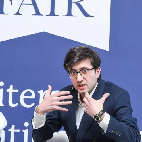 Professor David Maziashvili has dark brown hair, wearing a white shirt under a navy blue jacket. He's talking and gesturing with his hands. He's sitting down with a 'London Book Fair' backdrop behind him.