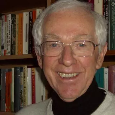 Alan has grey hair, glasses and is smiling at the camera. He's wearing a black shirt under a cream coloured jumper. He's standing in front of a bookcase.