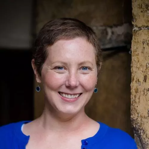 Dr LaPorte is leaning against a wall and smiling. She has cropped brown hair and is wearing a bright blue cardigan and blue earrings. 