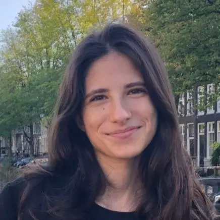 Nicoletta has long dark wavy hair and she is smiling at the camera. There are trees and buildings with large windows behind her. 