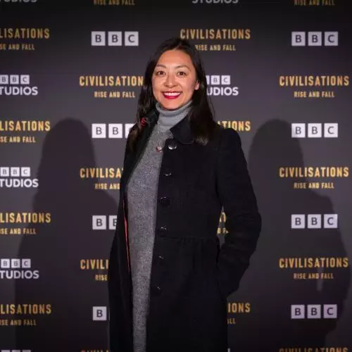 Jessica has long dark hair and is smiling at the camera. She's wearing a grey dress under a long black coat. She's standing against a 'BBC Civilisations: Rise and Fall' backdrop