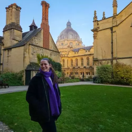 ellie has long dark brown hair worn in a pony tail. She's wearing a navy blue jacket with long royal blue scarf. She's standing in a college garden with college buildings and the Bodleian Library behind her.