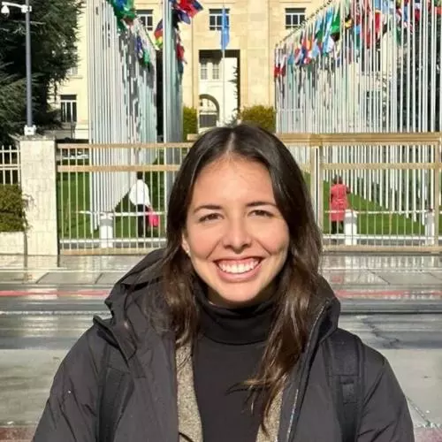 luciana has long dark brown hair and is smiling at the camera. She's wearing a black jumper and grey woollen cardigan under a grey anorak. She's standing in front of the country flags outside the UN Palais des Nations, Geneva.