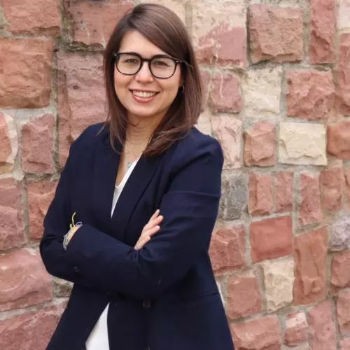 Maryhen had shoulder length brown hair, is wearing glasses and smiling at the camera. She's wearing a white top under a navy blue jacket. She's standing against a stone wall. 