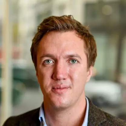Neil has brown hair, green eyes and is wearing a pale blue shirt under a brown woollen jacket.  