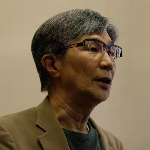 takehiko kariya has short grey hair, glasses and is wearing a grey top under a tan coloured jacket. 
