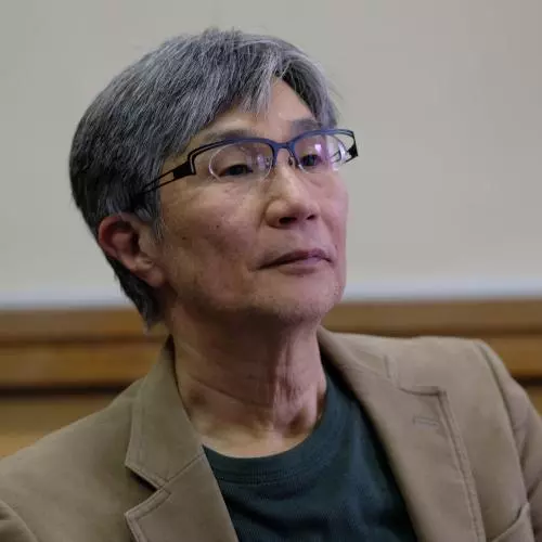 takehiko kariya has short grey hair, glasses and is wearing a grey top under a tan coloured jacket