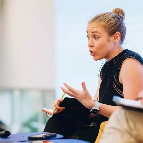 dr marnie howlett has blonde hair worn in a bun. She's wearing a black short sleaved dress and is talking whilst gesturing with her hands. 