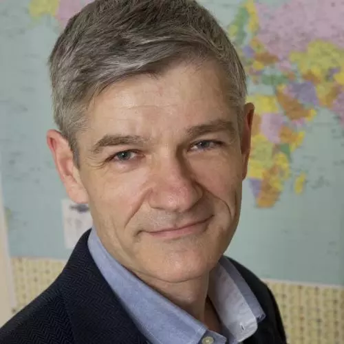 Roy Allison has short grey hair and is looking towards the camera. He's wearing a pale blue shirt with smart navy blue jacket. There is a world map behind him. 