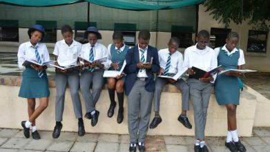 A row of school children in white and grey uniform sitting on a wall reading books. 